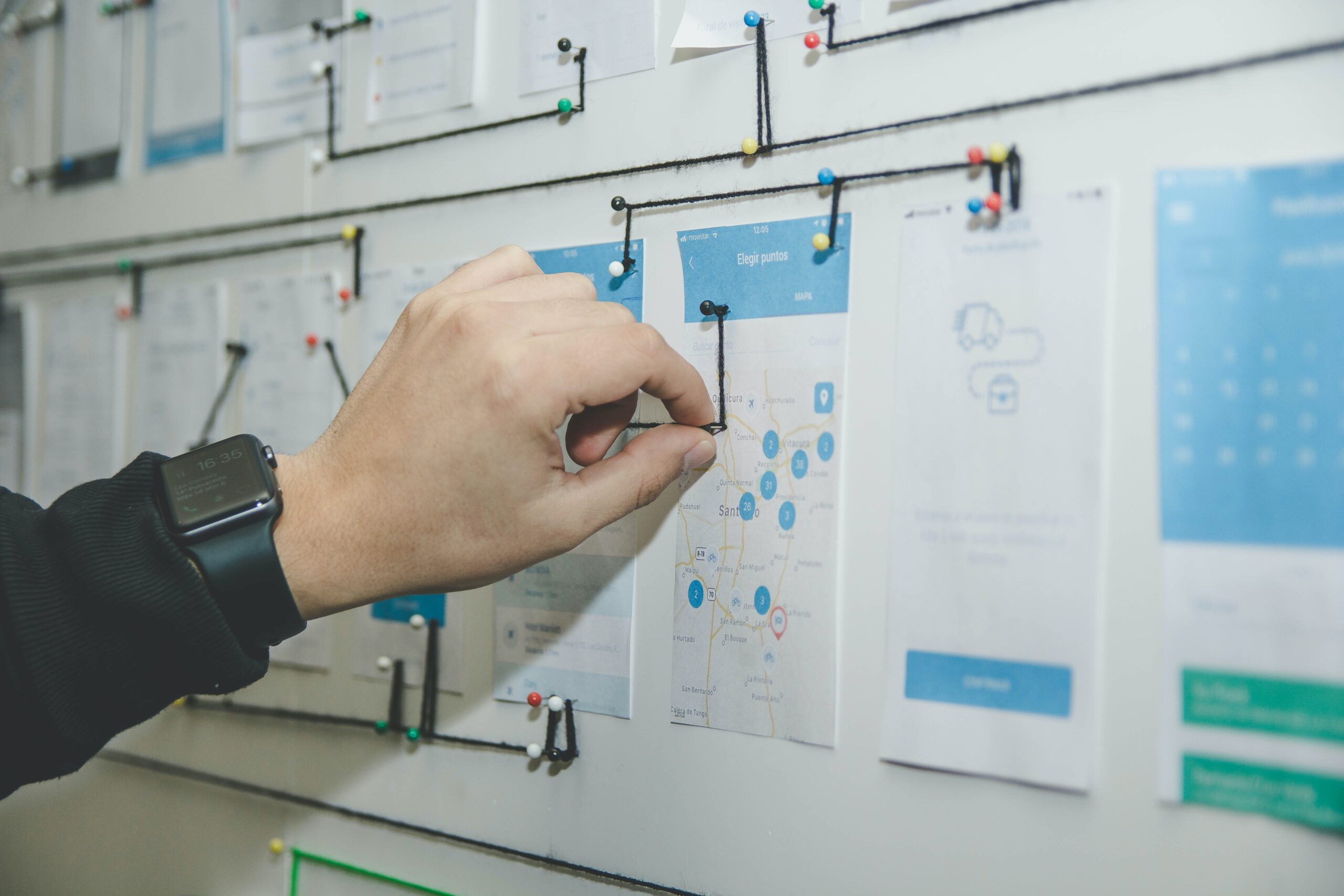 A hand wearing a smartwatch pins a string to documents and maps on a whiteboard, creating a network of connected papers with push pins and lines.
