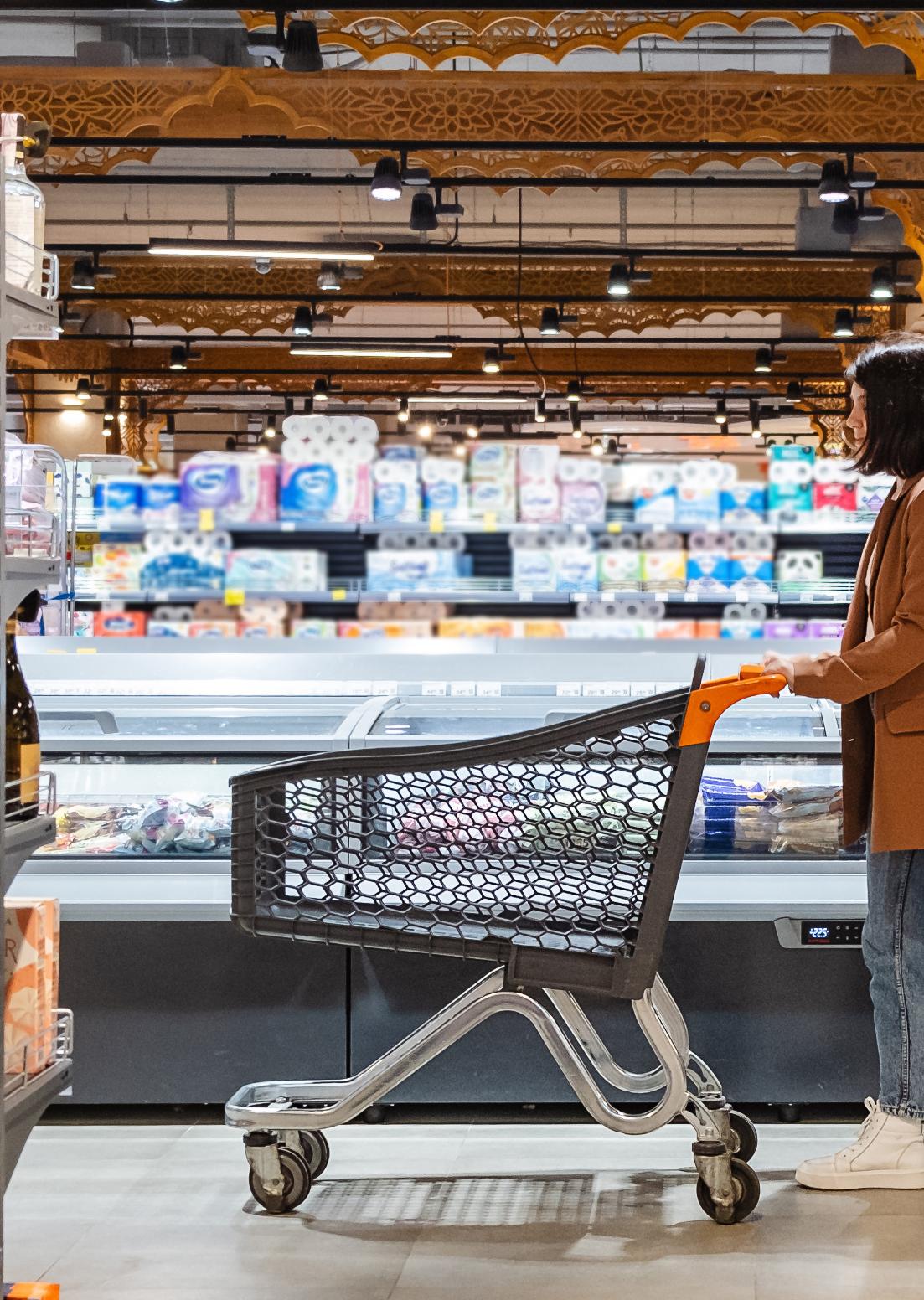 A person stands beside a shopping cart in a grocery store aisle, engaged in the process of selecting items from shelves stocked with various colorful products and refrigerated goods in the background.