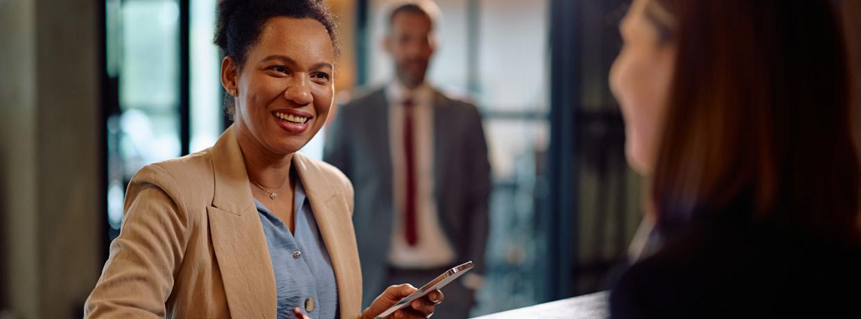 A smiling woman in a beige blazer holds a smartphone, embodying The Technology Pillar as she chats across a counter. In the background, a man in a suit stands slightly out of focus.