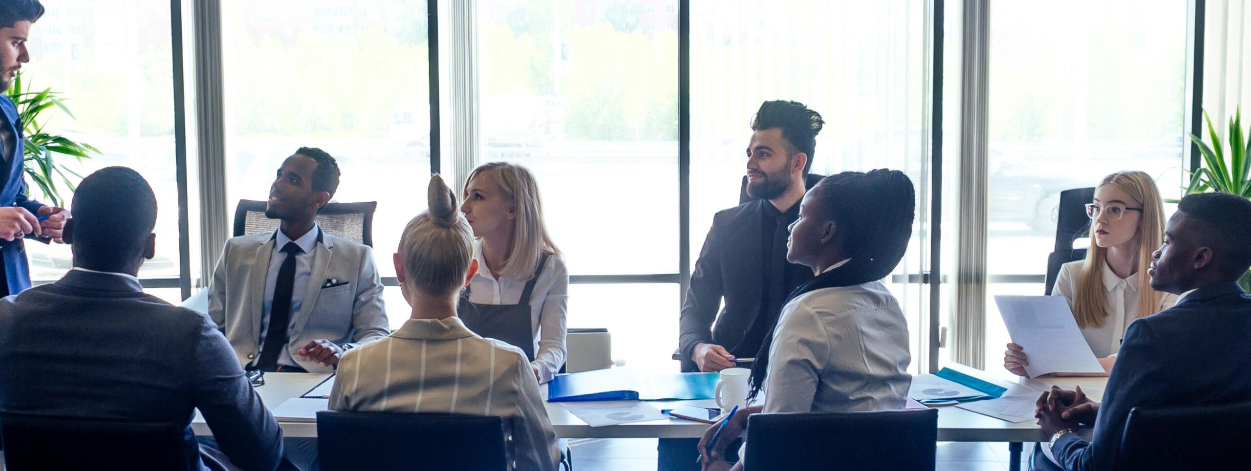 A diverse group of eight professionals sit around a conference table in a modern office, attentively listening to a standing colleague discussing The Technology Pillar. Large windows behind them let in natural light.