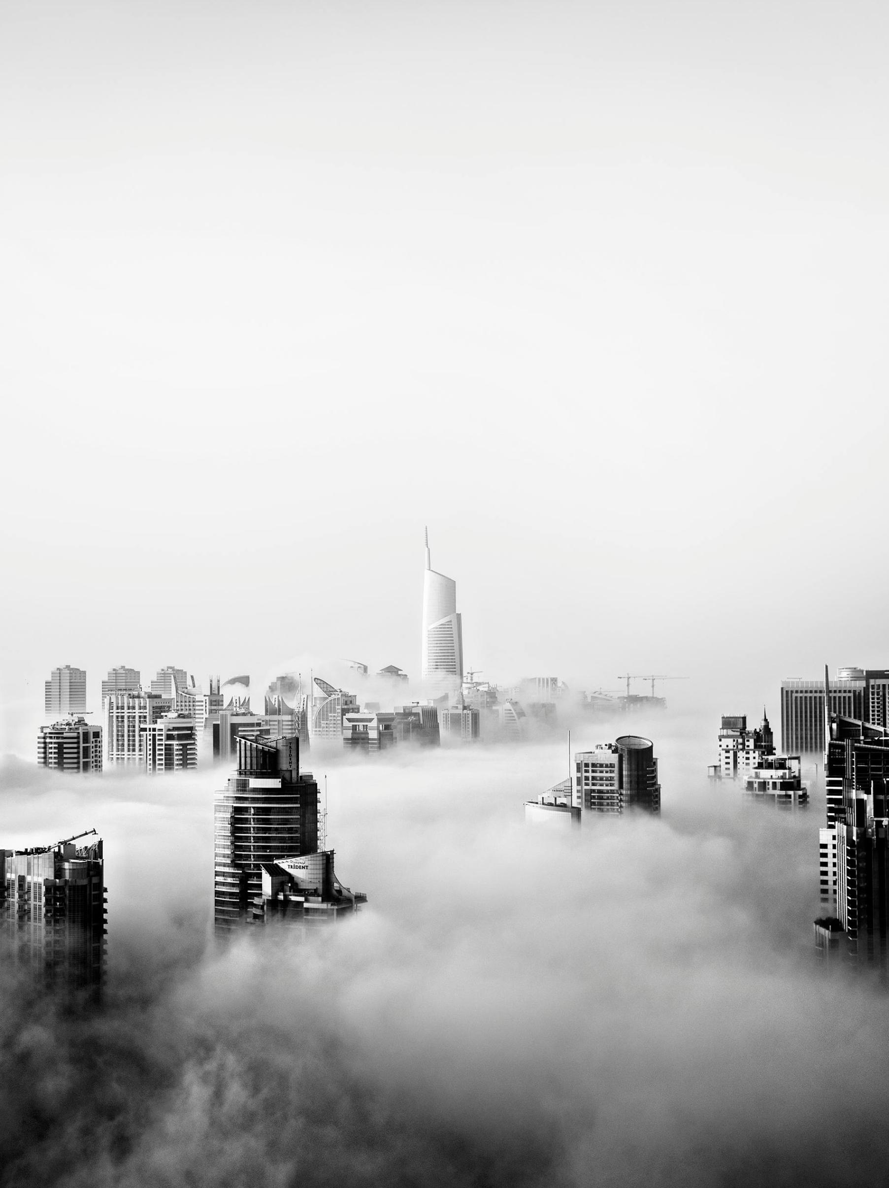 A city skyline with tall modern skyscrapers, partially obscured by dense fog, creates a dramatic and ethereal atmosphere. The tallest building—The People Pillar—stands centered, rising above the mist in this striking black and white image.
