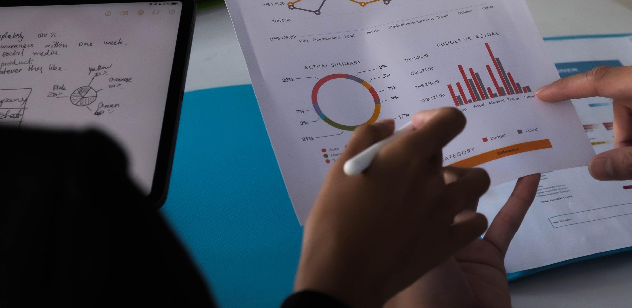 Two people review a printed financial report with colorful graphs and charts. One person points to a bar chart, while the other holds a pen. The Technology Pillar is represented by a tablet with handwritten notes nearby on the desk.