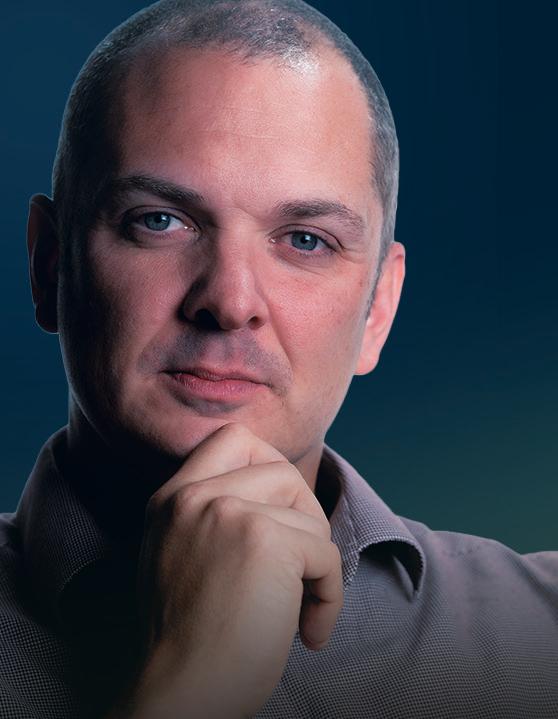A man with short hair and blue eyes looks thoughtfully at the camera, resting his chin on his hand. He is wearing a collared shirt against a dark blue background, embodying the reflective spirit of The People Pillar.