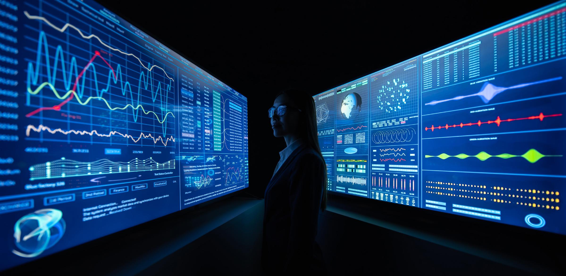 A person stands in a dark room surrounded by large, illuminated digital screens displaying complex data charts, graphs, and analytics in blue tones—an immersive scene powered by The Technology Pillar.
