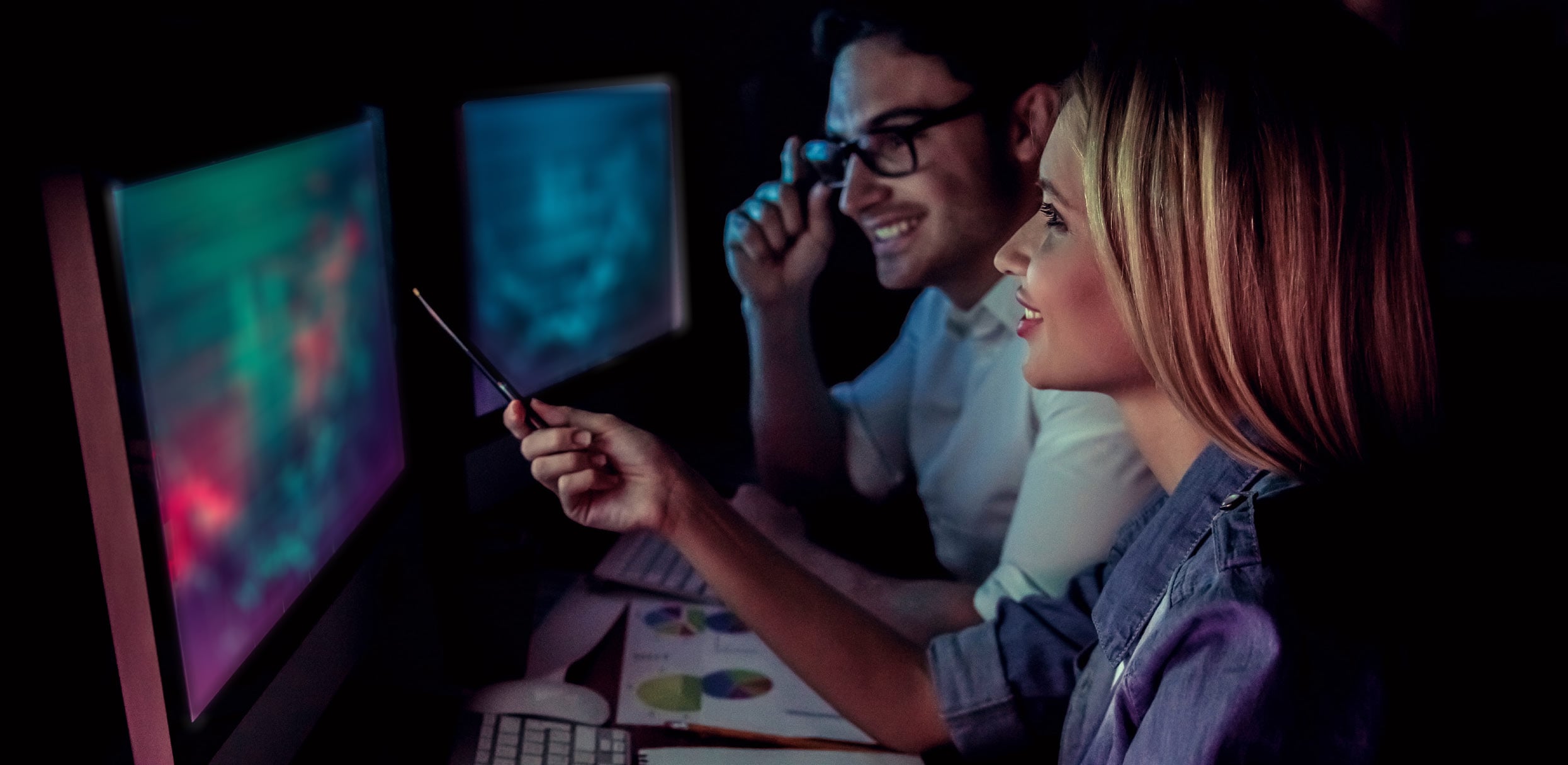 Two people sit in a dark room, looking at colorful graphics on computer screens. One person points at the screen with a pen as they discuss The Process, both appearing engaged and smiling. Papers with charts are spread on the desk.