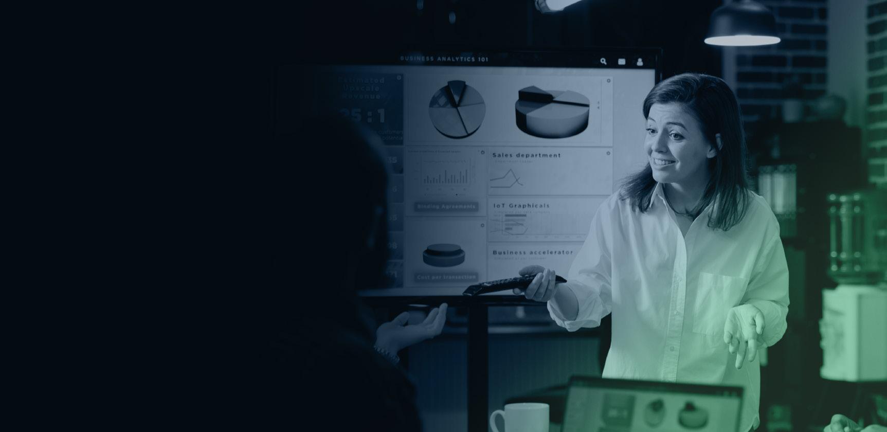 A woman presents data charts on a screen during a meeting in an office. She gestures while discussing The Process Pillar, with pie charts and graphs visible on the large display behind her.