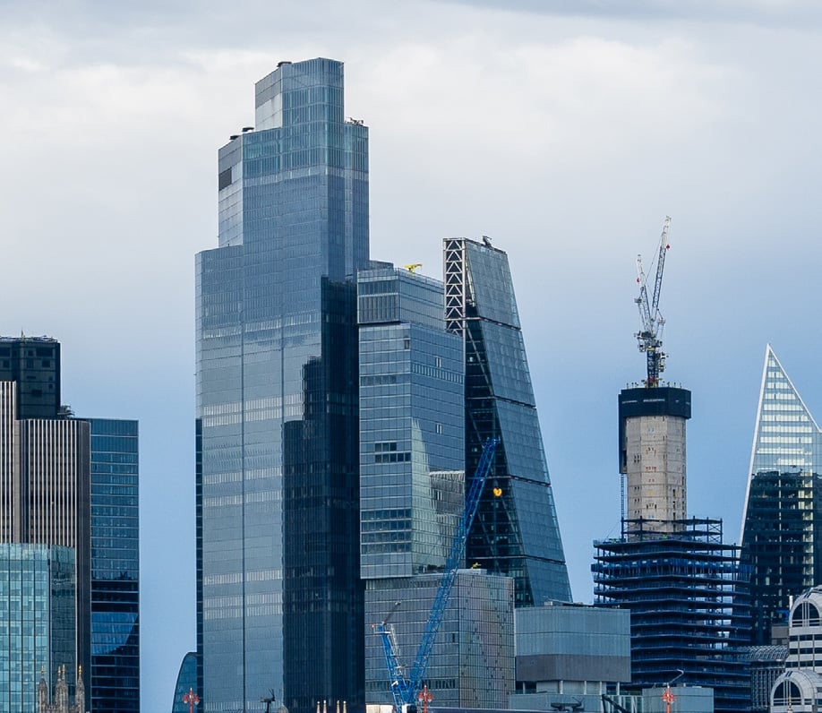 Several modern skyscrapers with glass facades stand tall in a city skyline under a cloudy sky. Construction cranes highlight the process of ongoing development transforming the urban landscape.