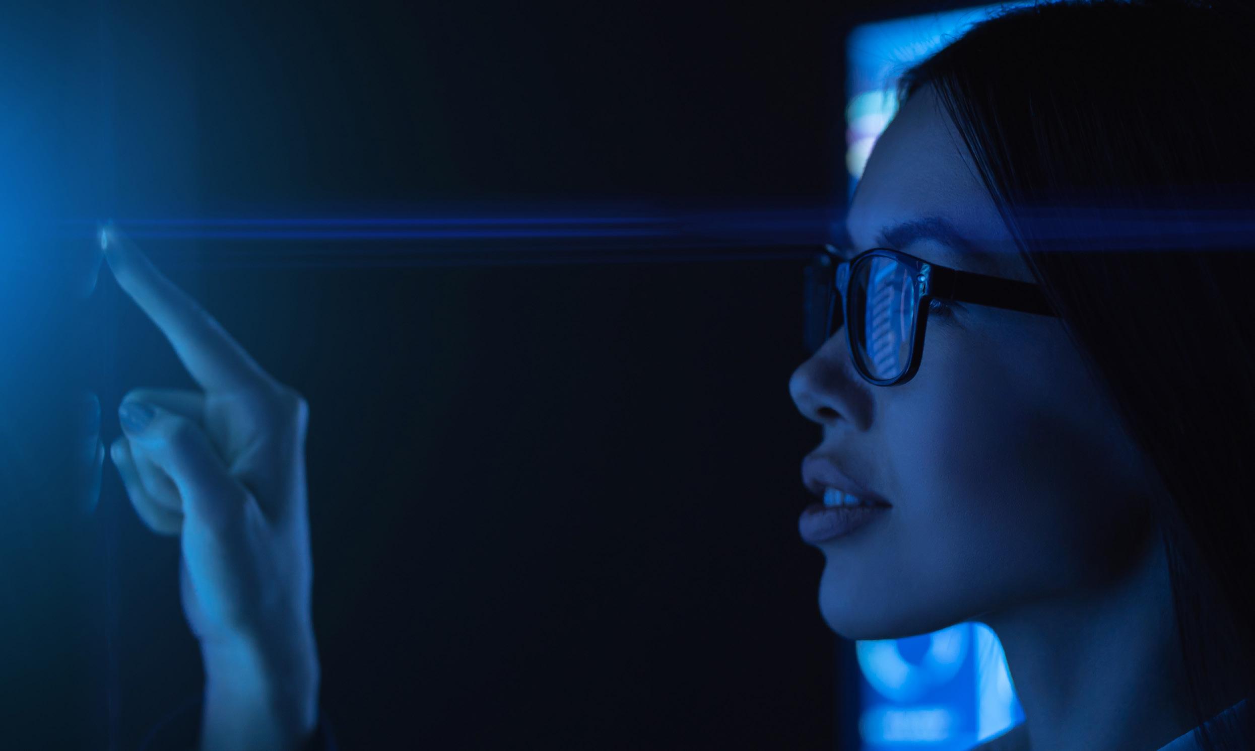 A woman wearing glasses touches a glowing screen in a dark room, illuminated by blue light from The Technology Pillar, with a digital display reflected in her lenses.
