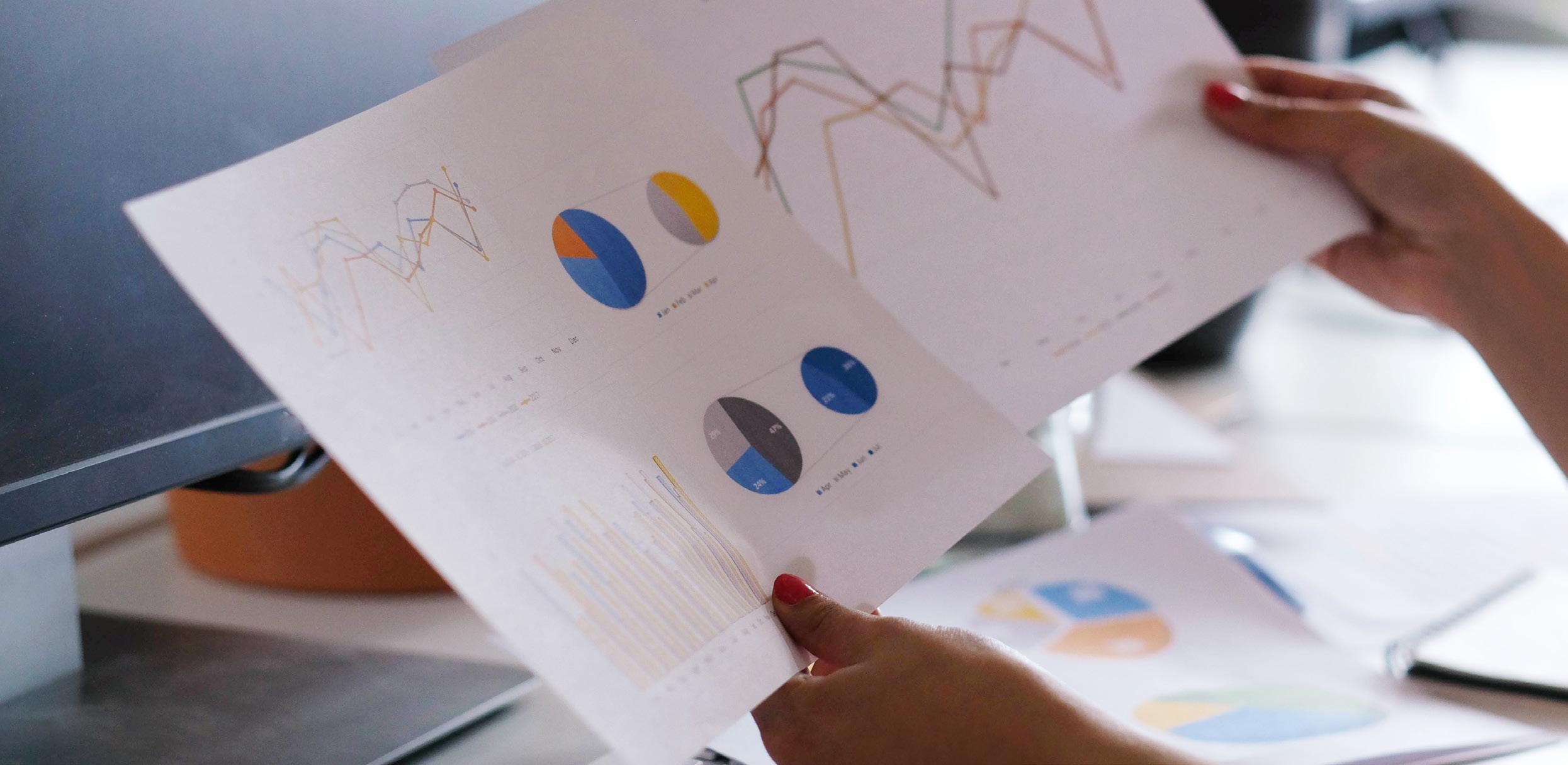 A person holds printed charts displaying line graphs and pie charts from The Process Pillar, analyzing colorful data visualizations at a desk in front of a computer monitor.